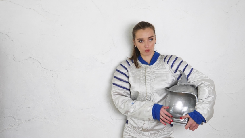 a girl in an astronaut costume holds a fantastic helmet in her hands stands against the background of a white wall and looks around