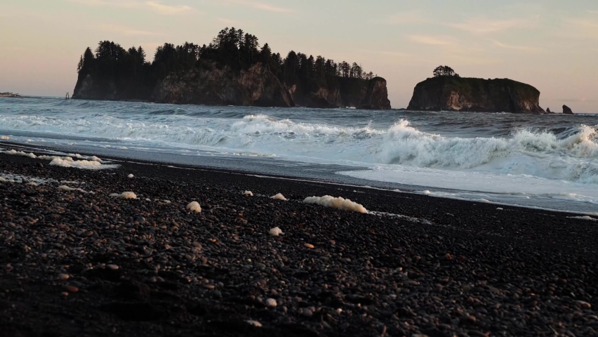 Gorgeous tilting up shot the pacific coastline with waves leaving behind sea foam, a beautiful small cliff island, and a colorful cloudy sky during sunset at the famous Ruby Beach in Forks, Washington