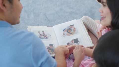 Asian family look at photographs on old picture album recalls the past. Young beautiful couple sitting with little kid daughters flipping pages of photo book and enjoy remembering relationship memory.