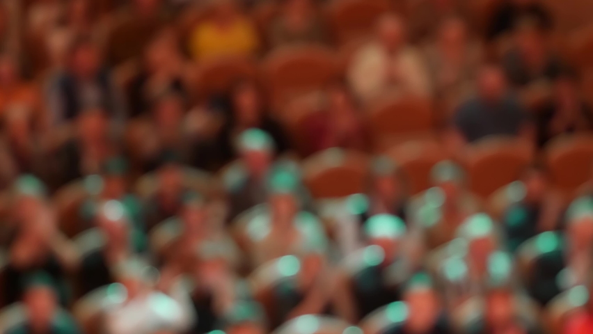 Unrecognizable out of focus spectators in hall intense applaud while sitting in armchairs. Visitors enjoy performance. Rock concert. Multi-colored moving spotlights create festive mood. Optical blur