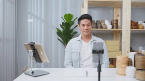 Asian handsome man sell vase product online live streaming at home. Young business male using laptop computer shows goods to customer and present detail. Remote buying and purchase shopping concept.
