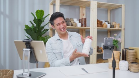 Asian handsome man sell vase product online live streaming at home. Young business male using laptop computer shows goods to customer and present detail. Remote buying and purchase shopping concept.

