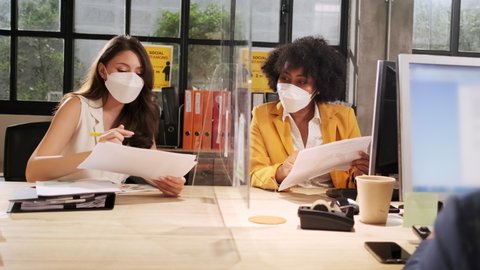 Two female coworkers team with face mask working in new normal office. COVID-19 protection by cleared partition, business workplace office, social distancing for pandemic health, disease prevention.