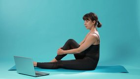 Fit woman doing stretching exercise and watching meditation video on laptop in studio. Athlete doing arms and legs stretch after workout exercise, following online yoga lesson on computer.