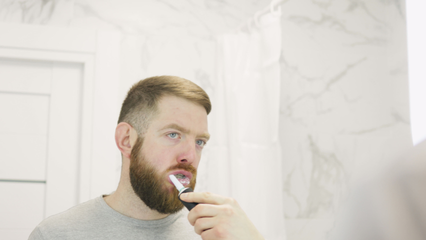 Bearded man brushes his teeth in front of mirror. Male using electric toothbrush and cleaning mouth doing morning oral dental care routine in bathroom.