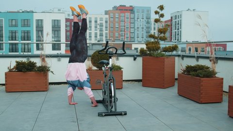 Athletic girl make acrobatic trick on bike. Caucasian woman exercising on stationary cycle machine bike on house rooftop gym. Modern sport recreational activity, workout, healthy lifestyle concept