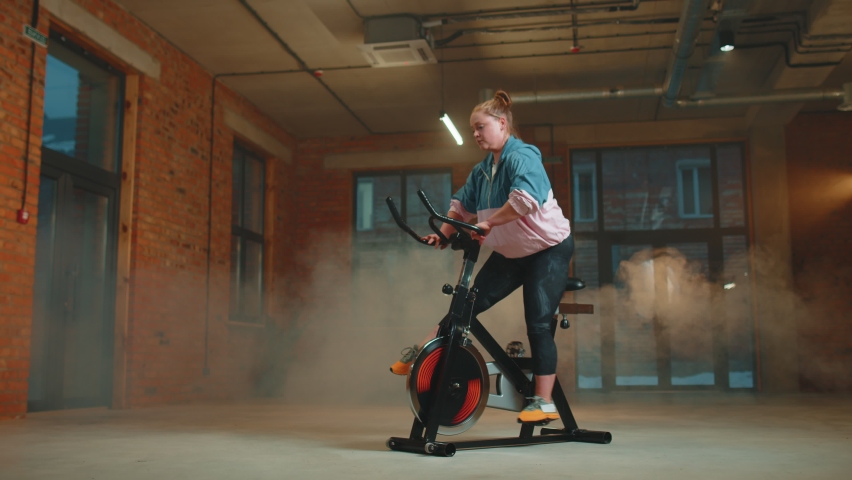 Athletic woman riding on spin stationary bike training routine in gym with haze. Fit girl making workout cardio training on exercise cycle bike indoors. Weight loss, fat burning. Modern sport Royalty-Free Stock Footage #1084863841