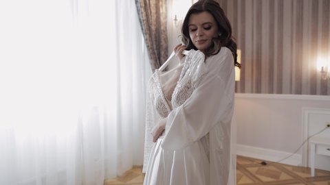 Bride in lingerie standing with her wedding dress. White boudoir dress. Morning preparations indoors. Luxury bride with hairstyle and makeup in a hotel or apartment. Woman in white night gown and veil