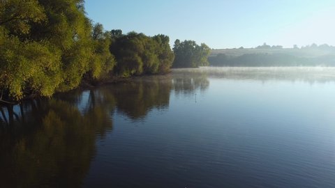 Pond with shore overgrown with willows and other trees, morning fog rising over the water at autumn, aerial view when moving forward above the water 
