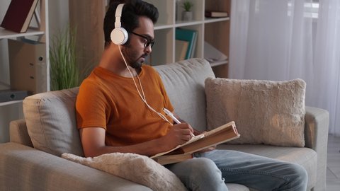 Study language. Smart man. Audio lesson. Handsome guy in headphones writing notebook sitting sofa with book light room interior.
