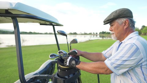 4K Portrait of Smiling Asian senior man golfer standing by golf cart on golf course at summer sunset. Healthy elderly male enjoy outdoor lifestyle activity sport golfing with friends at country club.