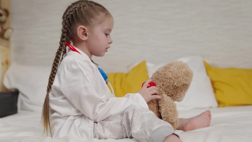 little kid plays with teddy bear sitting on bed in hospital, childhood dream of becoming doctor, kid veterinarian examines toy doll with stethoscope, pretend heartbeat, little daughter treats friend Royalty-Free Stock Footage #1084878088