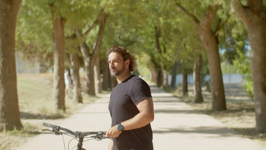 Front view of cheerful man standing on alley with bike in hands. Medium shot of happy muscular beadred man standing in park, looking at camera, devoting time to physical activity. Sport concept