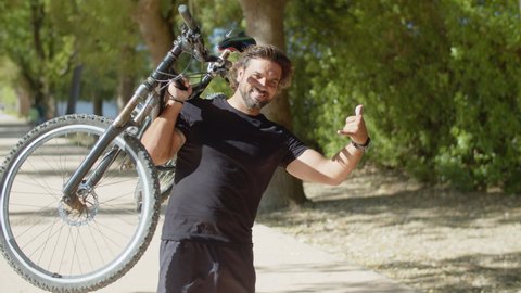 Man with disability lifting up bike and showing shaka sign. Medium shot of sportsman looking at camera, standing in summer park, celebrating goal achievement. Sport, disability, winning concept