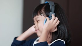 Video 4K Portrait of a Thai Asian kid girl aged 4 to 6 years old, cute face, holding headphones, she is listening to music in a relaxed manner. Happy and joyful, bright eyes, healthy.