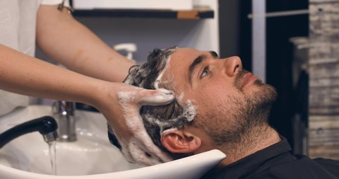 A handsome young man with a smile is resting in a hairdressing salon. After dyeing the hair brown, the hairdresser thoroughly washes the young man's head with water. Slow motion.