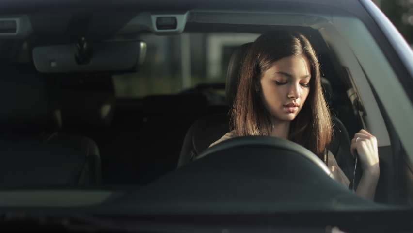 A young woman is sitting in a car in the driver's seat. She is putting on her seat belt and looking at the camera. She is smiling. 4K 50fps Royalty-Free Stock Footage #1084886215