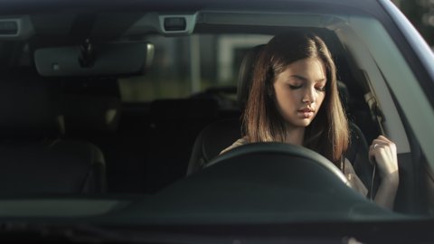 A young woman is sitting in a car in the driver's seat. She is putting on her seat belt and looking at the camera. She is smiling. 4K 50fps