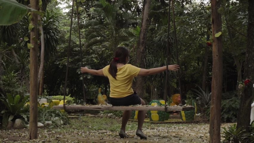 The back of a young Asian Female on an outdoor swing, her arms outstretched bracing herself as she rocks back and forth in a garden in Kanchanaburi, Thailand