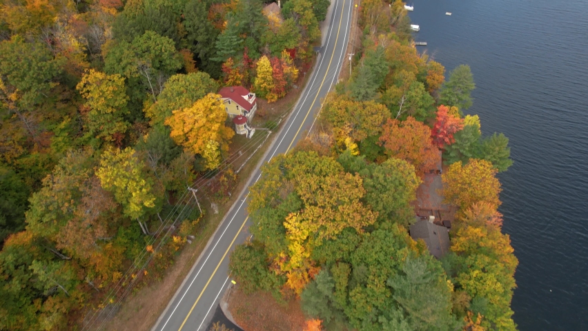 Aerial View of Dark Vehicle on Road in Colorful Autumn Landscape, Vivid Forest by Lake Sunapee, New Hampshire USA, Tilt Up Tracking Drone Shot