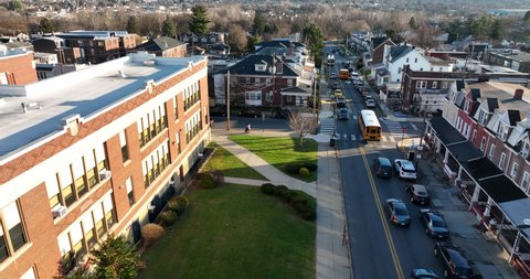 Aerial tracking shot of yellow school bus past public school building. Students live in urban city homes and houses. Bus stop in town.