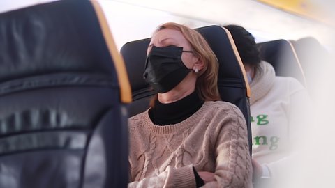 Young woman with face mask feeling unwell on plane, she is sitting with her eyes closed. Stress, headache, motion sickness and airsickness on plane during pandemic.
