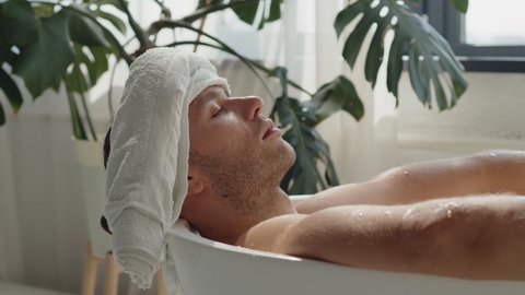 Pano shot of relaxed handsome caucasian young men lying in spa bath with white towel on his head, eyes closed. Relaxing spa treatments in eco bathroom. Mens modern wellness gain strength concept