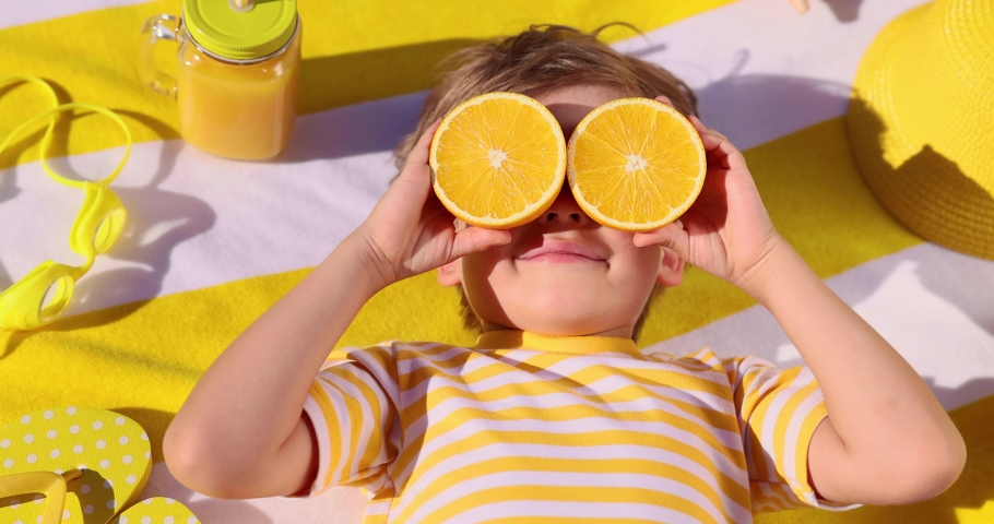 Happy child holding halves of orange fruit like a sunglasses. Funny kid lying on striped yellow towel. Boy taking sunbath outdoor. Healthy eating concept. Slow motion 4K | Shutterstock HD Video #1084894885