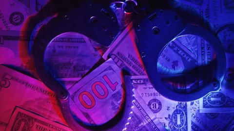 Close up of rotation metal handcuffs over stack of dollar banknotes. Concept of financial crimes or bribery and cash corruption police and deputies or fraud. Red and blue light from a police flashers.