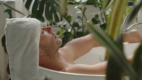 Pano shot of relaxed handsome caucasian young men lying in spa bath with a glass of champagne and white towel on his head, eyes closed. Relaxing treatments in eco bathroom. Mens wellness gain strength