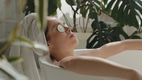 Pano shot of relaxed bautiful caucasian young woman lying in spa bath with cucumber on eyes and white towel on head, eyes closed. Relaxing spa wellness treatments in eco design bathroom