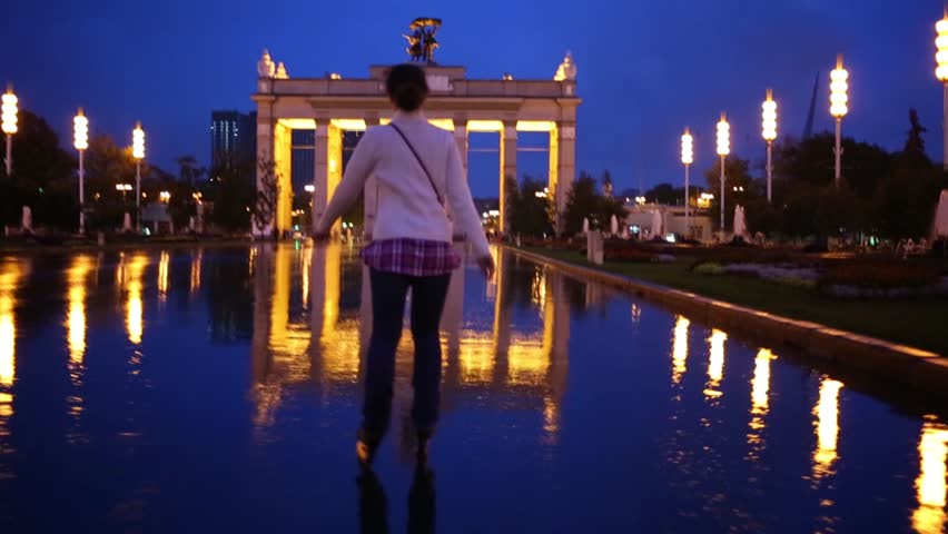 Young woman rides roller skates through puddles under the rain at evening park towards entrance arch.