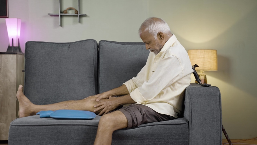 senior man using Hot water massage bag for knee joint pain relief while sitting on sofa at home - concept of natural hot water treatment, muscle relaxtion and osteoarthritis.