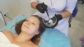 Cosmetologist applies mask to skin of woman's face for therapeutic purposes. Patient is lying on couch and thick transparent substance is applied to face with brush. Rejuvenation, acne treatment