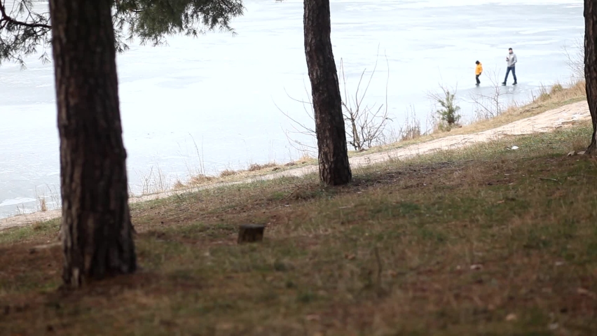 A male traveler walks along the river bank along a forest path in winter. On background dad and son are walking on a frozen lake. Danger of walking on ice.