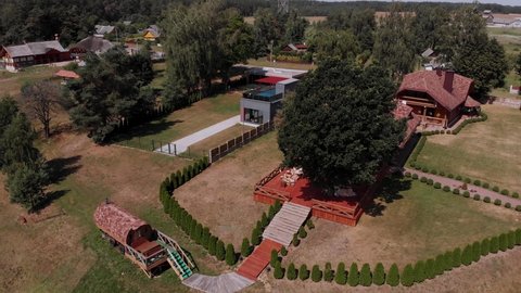 Aerial summer view of large green oak growing on wooden red terrace of house surrounded by thujas. Modern village with old huts and modern cottages. Bathhouse is located near a large staircase.