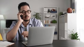 Feeling happy lively positive and energetic. Young Asian businessman wearing glasses talking on phone sitting at desk with laptop. slow motion