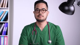 Portrait shot of young Asian male doctor in surgical uniform listening talking explaining and discussing with patient online, making video call, looking at camera.