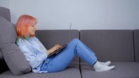 Young white woman using internet technology for networking and doing distant work on lockdown. Girl with dyed hair typing on laptop computer while lying on couch at home. Freelance work concept in 4k