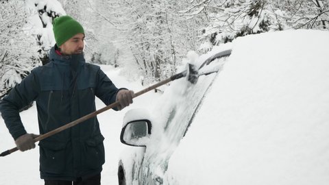 Man cleaning snow-covered car with brush after heavy snowfall. Snowstorm, bad winter weather, natural disaster. Person cleans side windows of his car from snow.