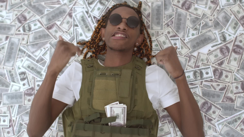 African-American man in bulletproof vest is lying on pile money and dancing, money rain. Black rapper is lying in pile of dollars bills are falling on it. Concept rich Royalty-Free Stock Footage #1084933213