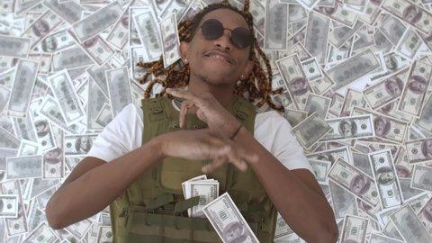 African-American man in bulletproof vest is lying on pile money and dancing, money rain. Black rapper is lying in pile of dollars bills are falling on it. Concept rich