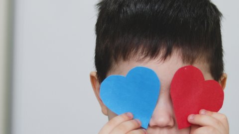 close up video of a happy kid playing with two paper heart at the eyes. blue and red heart, concept of valentine's day. love. brunette hair boy.