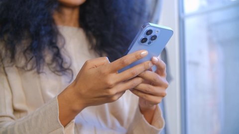 Young adult african woman using modern mobile phone at home on lockdown.Black female browsing internet and new feed on social media app in trendy smartphone with triple camera. Mobile internet concept