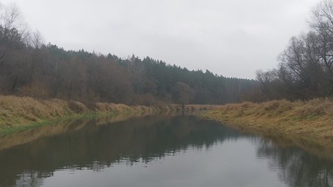 The boat drifts slowly down an old river bed, with tall grass, bushes, and trees growing along its banks. The forest is reflected in the calm water. The weather in November is overcast.