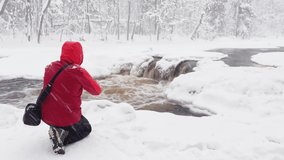The photographer does to a photo and video the wild nature at snow storm by smartphone, he is dressed in red color jacket, the wild frozen small river in the winter wood, ice, snow-covered trees