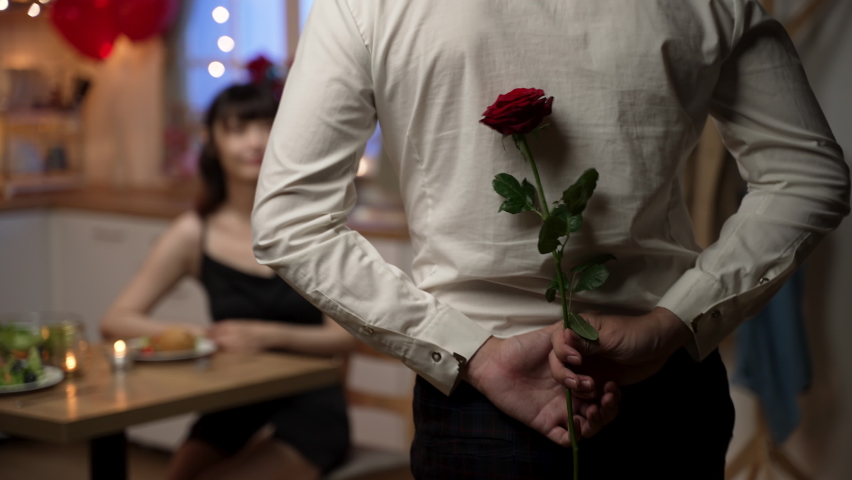 rear view of asian man wearing suit walking toward girlfriend with rose behind. boyfriend kneeling down to give lover a flower on valentine’s day dinner date at home