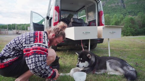 Male Camper Pours Dog Food Into A Plastic Bowl For His Pet Alaskan Malamute. full shot