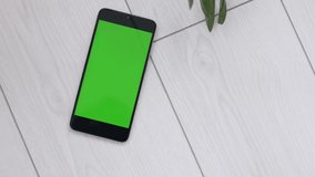 Closeup top view 4k video footage of modern smartphone laying on wooden white vintage background indoors. Cute leaves of money tree flowers laying near gadget with empty blank green screen.