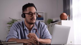 Feeling satisfied and positive at work. Young Asian businessman wearing headset on video call with clients on laptop. Young Asian man in glasses giving online educational class lecture